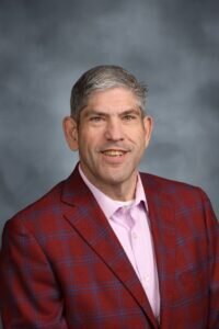 Headshot of a middle-aged man with short, graying hair. He is smiling and wearing a light pink button-down shirt under a red and blue plaid blazer against a mottled gray studio background.