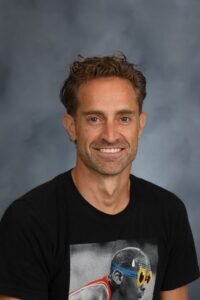 A man with short, curly brown hair smiles at the camera. He is wearing a black t-shirt with an image of an athlete in goggles on it. The background is gray and softly blurred.