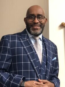 A man with glasses and a beard smiles, wearing a blue plaid suit, light blue checkered shirt, and a silver tie. He stands indoors against a neutral background.